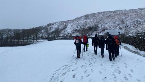 Six people are trekking through a snow covered field in warm clothing. The is a snow covered mountain in the distance.