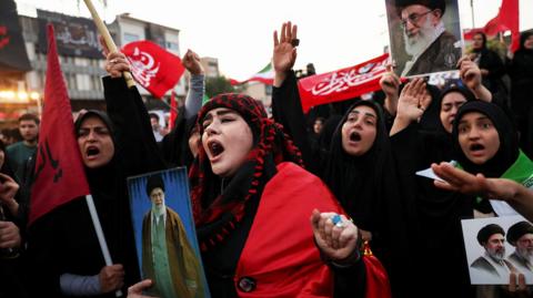 Woman holds picture of former supreme leader amongst crowd of people. All of them are shouting, with a number raising red flags in the air