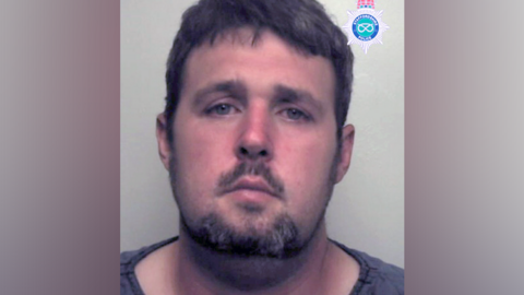 A mugshot of a man wit short black hair, a beard and a grey or black top.