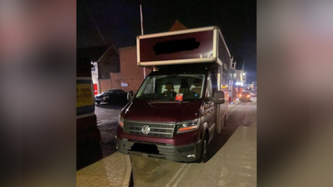 A large maroon VW van blocks the entrance/exit to Saffron Walden fire station.  It is dark. Lights from buildings and shops can be seen in the background. 