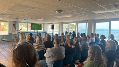 A group of people sat in blue chairs listening to a speaker at the front of the room. There is a green presentation to the right of the speaker at the front, who is holding a microphone. The room has large windows with a sea view.