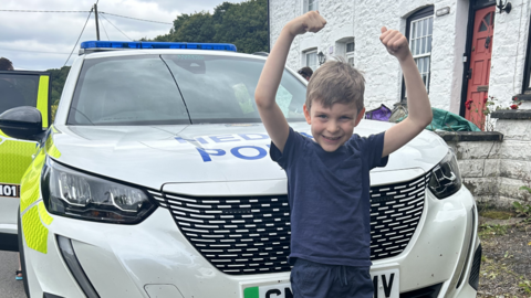 Macsen stood in front of a police car with his arms in a cheering gestur ein the air