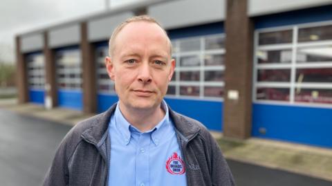A man with short fair hair, wearing an open blue fleece and blue shirt, stood in front of four garage doors housing fire engines. The man has a logo on his shirt, which reads "Fire Brigades Union".