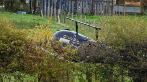 A black helicopter on its side in a field, partly concealed behind some bushes.