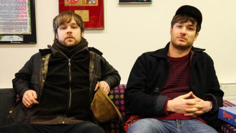 James Skelly and Nick Power sit side by side on a sofa. Skelly holds a corduroy hat in one hand and Power wears a cap and has his hands clasped. 