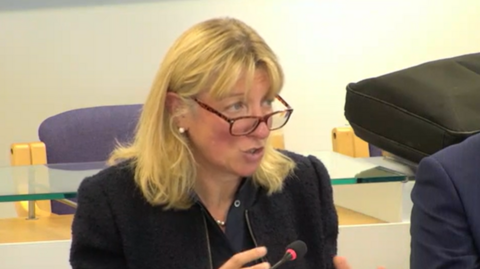 Jo Shiner has blond hair and glasses and is wearing a black suit. She is sitting down and speaking, answering questions from a panel of councillors in a council chamber