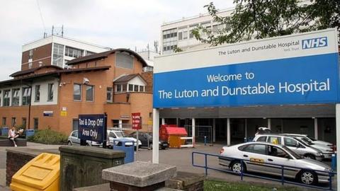 The exterior of the hospital with a large blue and white sign saying "Welcome to The Luton and Dunstable Hospital". There are silver taxis lined up and a black sign saying "Taxi & Drop Off Area Only. A four storey building is seen to the left of and also behind the sign.