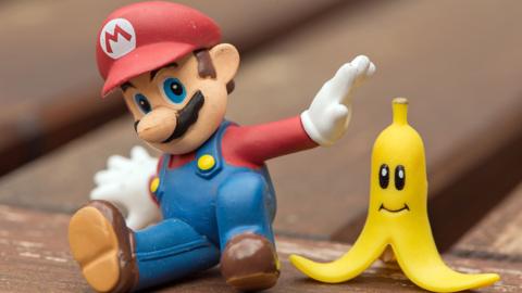 A small figurine of the animated character Super Mario, dressed in a red hat and blue overalls, sits on a wooden surface beside a yellow smiling banana-shaped toy.