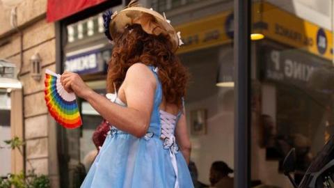 A drag performer is in a street. They're wearing a curly auburn wig and a blue dress, that is laced up at the back and holding a rainbow coloured fan. 