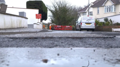 A wide shot of a road closed for resurfacing. There is a van parked on one side of the road, and a large red 'road closed' sign.