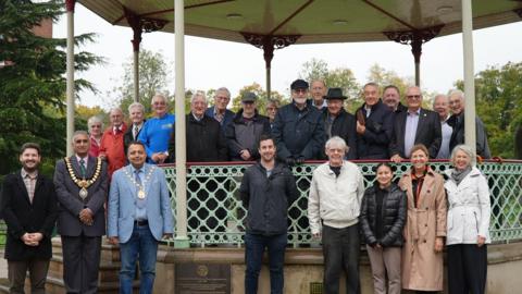 A group of people standing inside a bandstand smiling at the camera. They are stood alongside a plaque, installed on the base of the structure. 