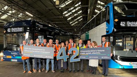 One single deck and one double decker bus inside a large open building (the depot). Several Stagecoach West workers stand before the buses, holding two large blue numbers that read 42. They are also holding signs with one saying "£20.5m investment in Gloucestershire's transport" and the other saying "Gloucestershire's future: clean, quiet, electric".