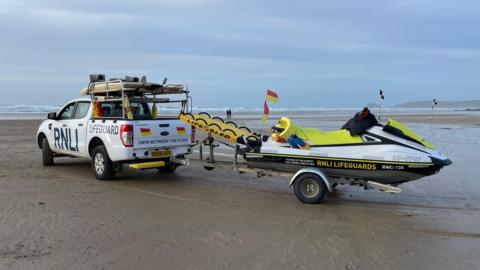RNLI Lifeguard car, with a RNLI lifeguard jetski attached to the back. It is on the beach. The sand is wet and brown. There are waves in the background. There are red and yellow flags and black and white flags in the background. The sky is blue and cloudy. 