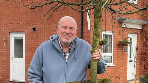 Jim Dann, a man who is standing on Jessup Road in Watton next to a tree. He has his hand around the tree and is looking directly at the camera. He is wearing a red and blue shirt with a blue zip-up hoodie over the top. 