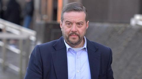 A man with grey hair and a glum expression. He has a moustache and beard and is wearing a light blue shirt and navy jacket.