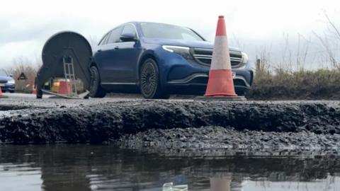 A pothole is photographed from ground level. Water can be seen in the crater, with large edges, and a car can be seen in the background. 