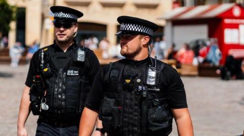 Two uniformed police officers are walking through a town. It is a sunny day and they are wearing hats and sunglasses. People can be seen blurred in the background