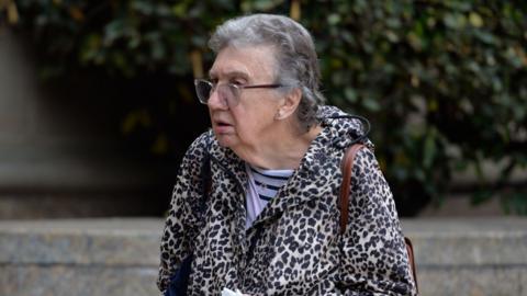 Patricia Robertson, a 77-year-old woman with short grey hair and clear-rimmed glasses, is walking right to left. She is wearing a leopard skin hooded jacket and a  light and dark blue striped shirt underneath. She has a brown leather bag slung over her shoulder.