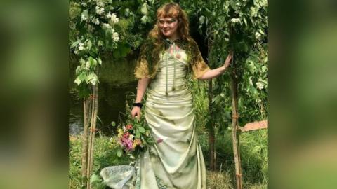 Megan Ruth-Trump stands beneath a floral arch beside a river. She is wearing a wedding dress and holding a bouquet of flowers in one hand