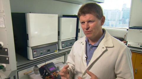 A man wearing a white overall and a blue shirt stood in a room with scientific equipment and a window behind him