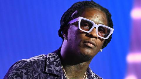 Young Thug with his hair tied back, wearing a black and white paisley shirt, white glasses and diamond earrings