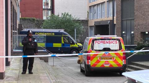 A police cordon at the foot of a block of flats. A uniformed police officer guards the steps. Two vans marked "forensics" are parked nearby. 