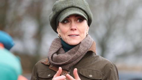 The Princess of Wales in a brown raincoat and wool scarf, wearing a green polo neck underneath. She is look to the side in thought and has her hands in front of her. Her brown hair is swept under a green tweed cap.
