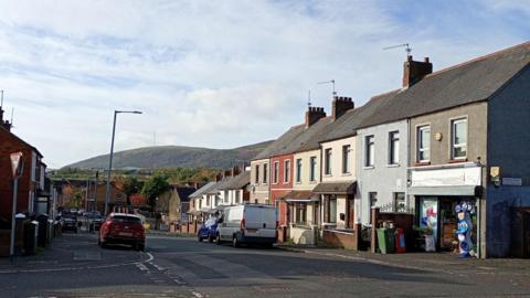 A Belfast street with houses on either side. There is a shop front on the nearest house on the right. There are a number of vehicles parked by the street. 