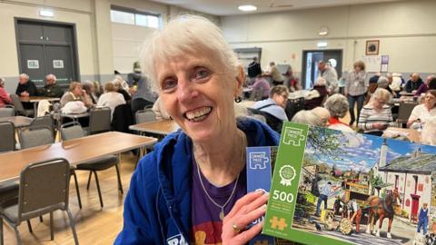 Lynn Pearl has short white hair, she is wearing a blue fleece with a purple shirt underneath and a silver chain with a pendant on it is around her neck, she is holding a couple of jigsaw puzzle boxes, behind her are people sitting chatting in a village hall. 