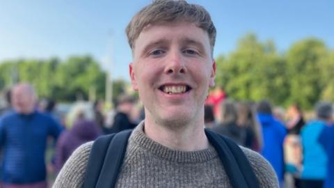 A man smiles into the camera. He has fair hair and is wearing a grey jumper. He has black backpack straps over his shoulders.