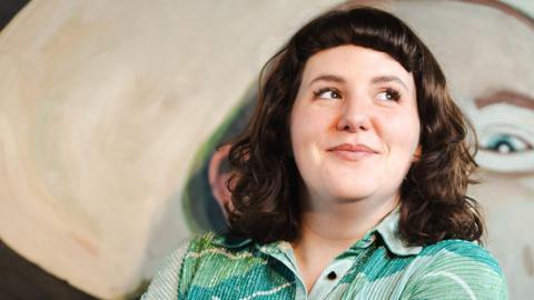 Amanda Dwyer standing in front of a large abstract painting with soft, rounded shapes and muted colours, wearing a patterned green shir