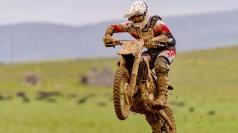 A motocross rider covered in mud as his bike is doing what appears to be a wheelie