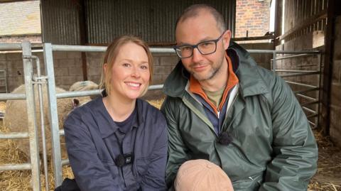 Laura and Tim are sat in front of their herd of sheep. Laura has dark blonde hair, tied back in a ponytail and wears a blue overall. Tim has a green jacket on over a blue top and wears blue-rimmed glasses.