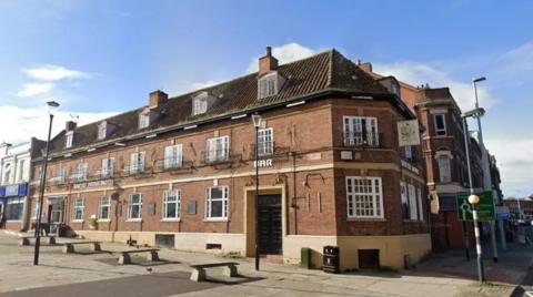 The Sneyd Arms Hotel, a three-storey red brick building with a series of dorma rooms in the top floor. There are benches situated in front of it, and a zebra crossing to the right hand side.