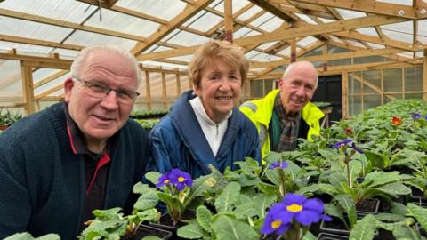 Adrian Thompson, left, Joy Baldry and Glen Unstead, in a high visibility jacket, stand near rows of polyanthus in a timber-framed greenhouse. Mr Thompson and Mrs Baldry are wearing blue jackets.