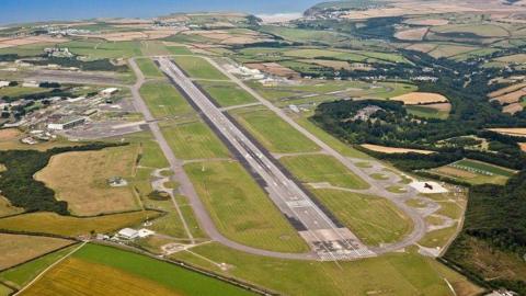 Aerial view of a plane runway. It is surrounded by green fields. The sea is in the background.
