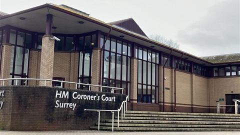 The exterior of a brick and glass building. A sign on the wall near entrance steps reads HM Coroner's Court Surrey.