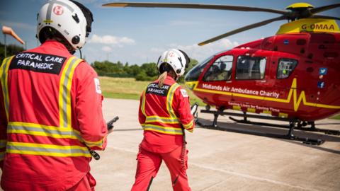 Two Midlands Air Ambulance crew walking towards a red helicopter. They are dressed in a res uniform with the words "critical care paramedic" on the back.