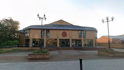 A modern, multi-story brick building with large windows and a central entrance. Two lampposts with multiple lights stand on either side of a paved area leading to the building.
