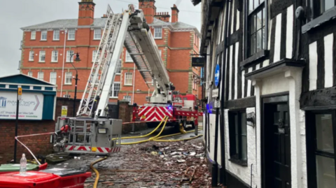A fire appliance with a cherry-picker is parked on a street covered in debris, in front of a black-and-white half-timbered building. 