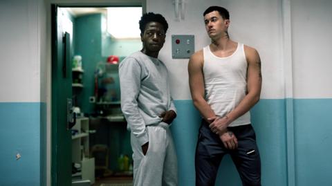 Two men stand next to a prison cell. The man on the left is wearing a grey tracksuit and is looking off camera. The man on the right is looking at the man on the left, and is wearing a white vest and tracksuit bottoms.