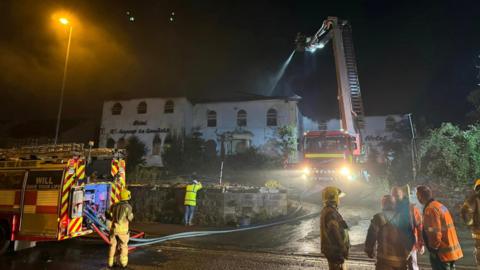 An aerial ladder platform is used to spray water on to the roof of the former La Gondola restaurant and hotel building, while firefighters stand in the foreground