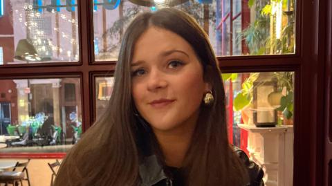 Stella Trimble looks into camera. She has a gold ear-ring and dark brown long hair. She is sitting in front of wooden framed window. Plants and candles can be seen behind with some reflective light shining back from one window pane.