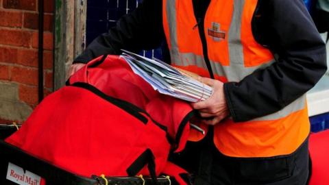 A postman photographed from the neck down holding a red sack of mail
