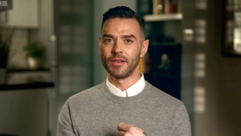 Matt Willis sits facing the camera - he has short black hair and wears a grey jumper
