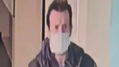 A CCTV image of Dean Hesketh, wearing a facemask and brown coat. It is of poor quality.