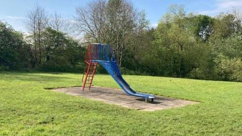 A blue and red slide sits on concrete. It is surrounded by grass and trees on a sunny day. 