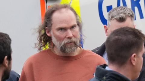 A man with long greying hair and a grey beard stands outside a garda van - he wears a red top