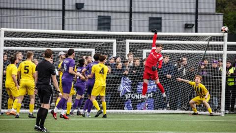 A group of footballers gathered around a goal as a ball comes flying in from the right. The goalkeeper, dressed in a red strip, is leaping into the air and holding the crossbar with one hand.