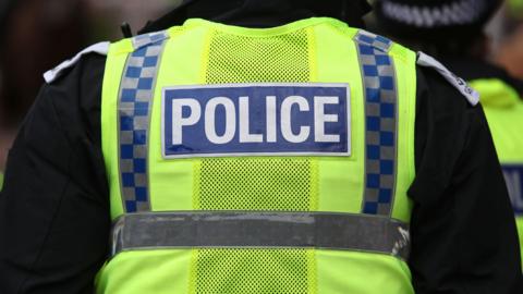 A police officer wearing yellow high vis vest with Police written on it, seen from behind